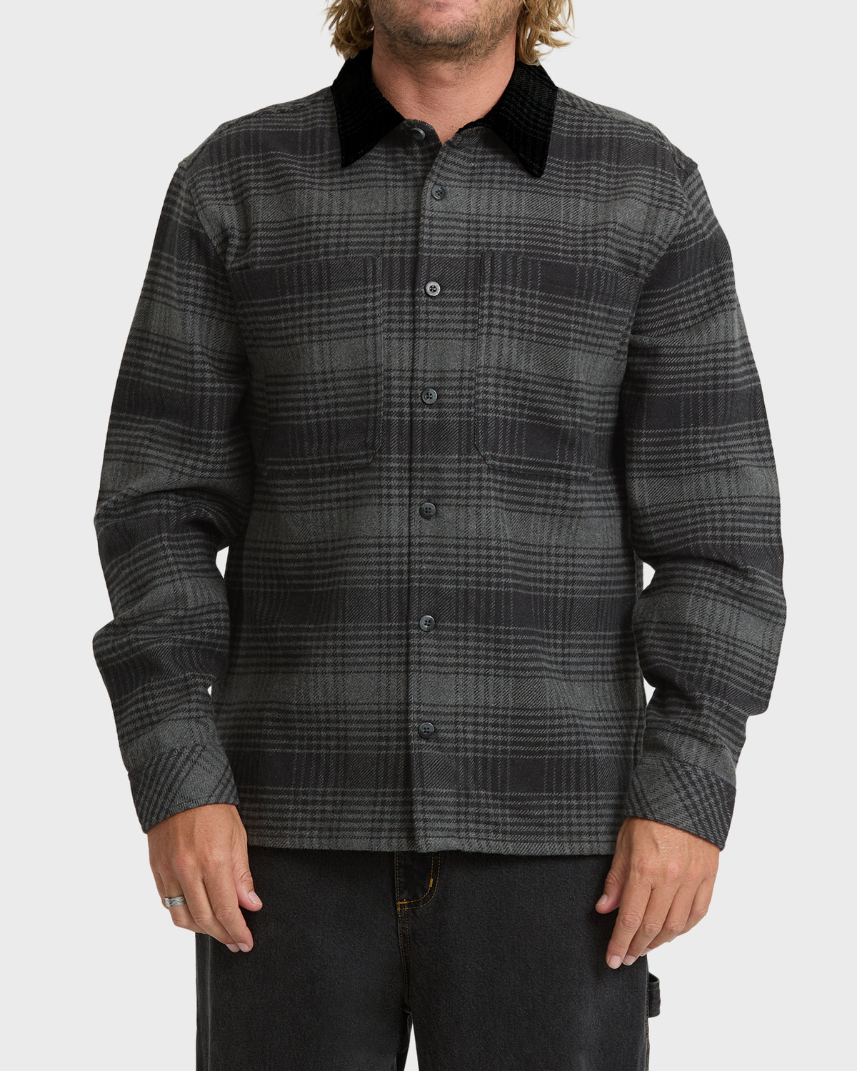 
A man wears a buttoned gray and black plaid long sleeve shirt with a black corduroy collar and two chest pockets.