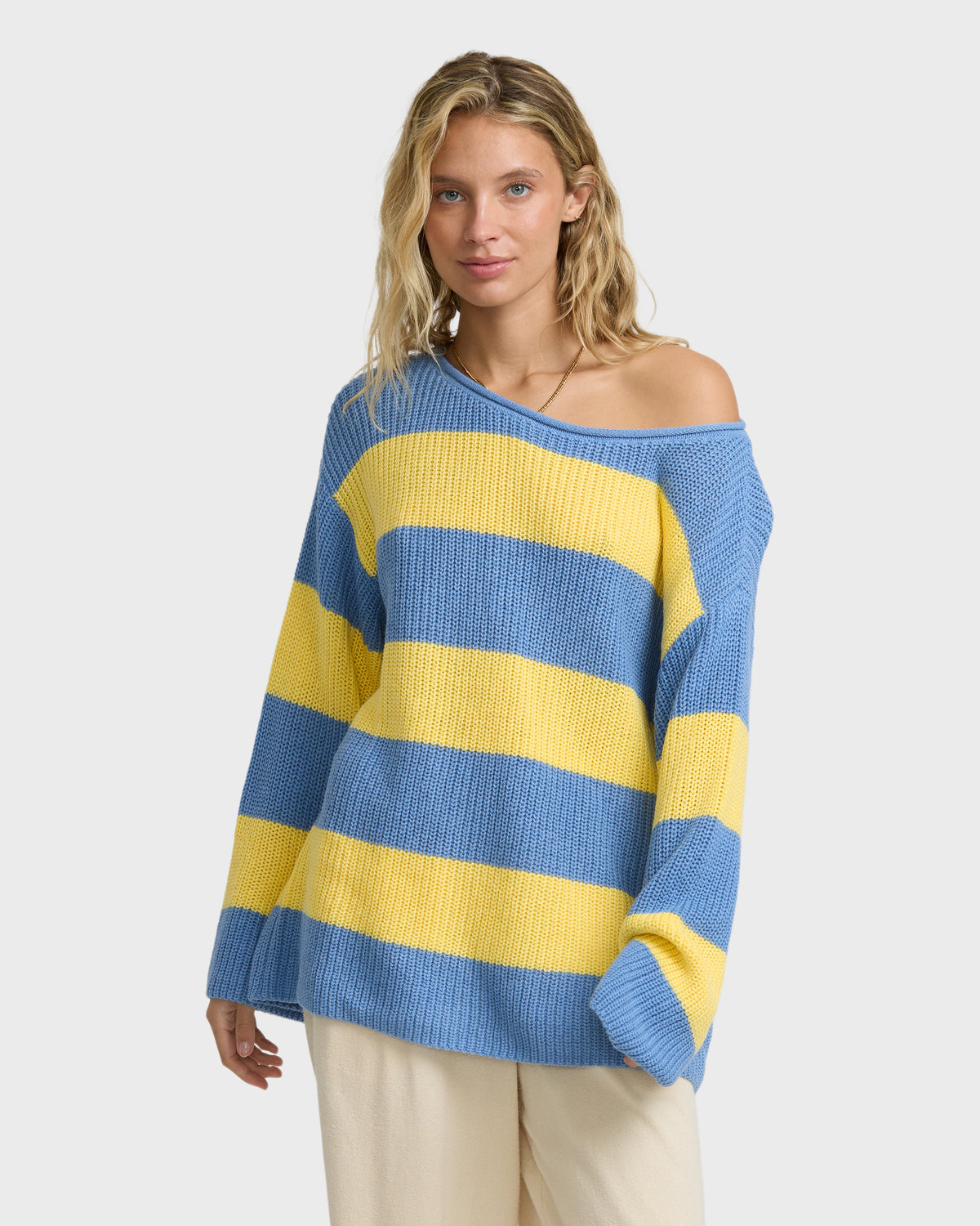 Blue and yellow striped off-shoulder sweater with ribbed texture worn with cream-colored pants.