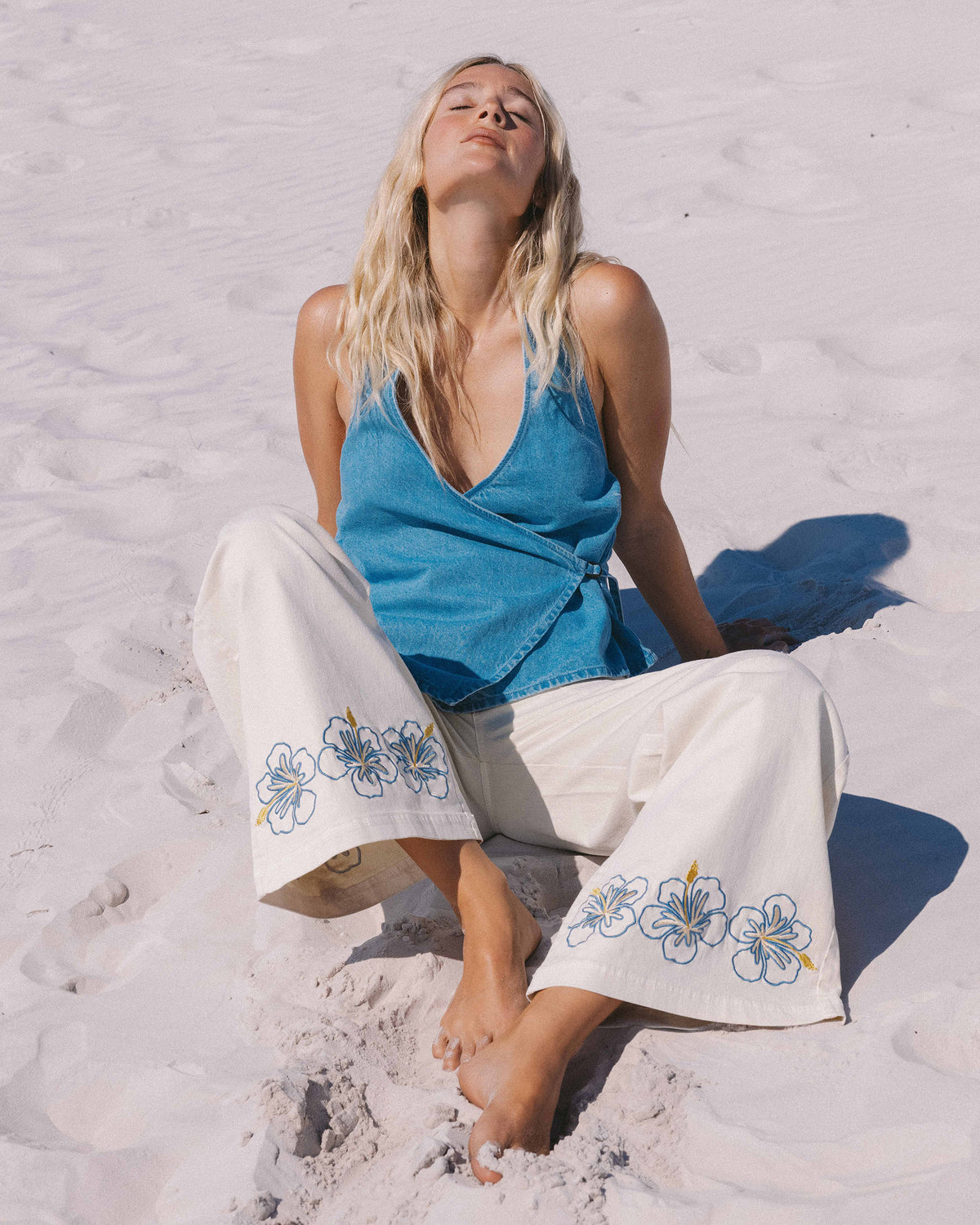 White pants with blue hibiscus flower embroidery on legs, displayed on model sitting on beach.