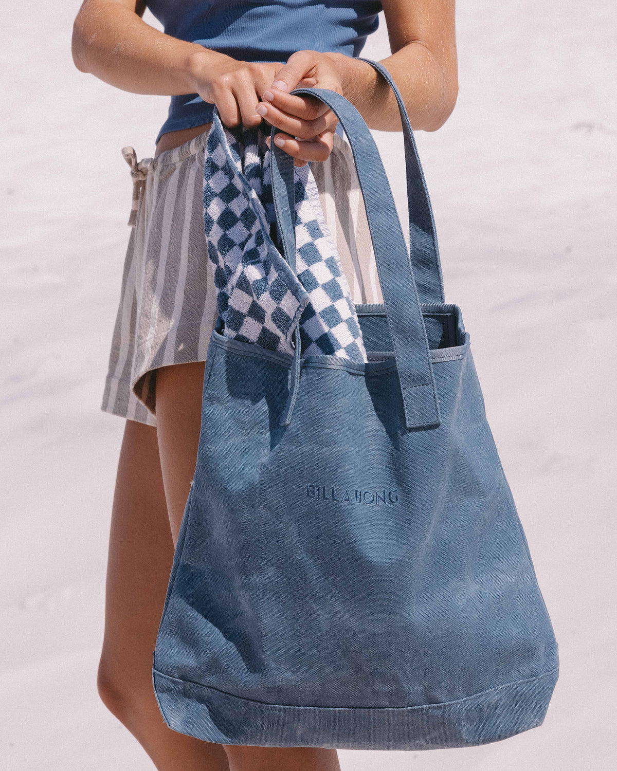Blue-gray canvas tote bag with shoulder straps, held by person wearing blue shirt and patterned skirt.
