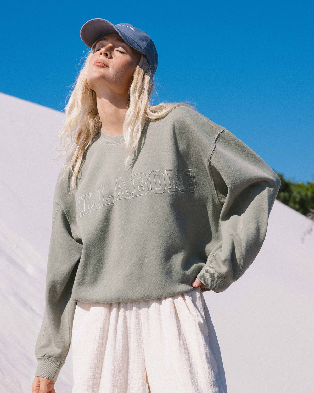 Sage green crewneck sweatshirt with embroidered text across the chest, worn with a baseball cap.