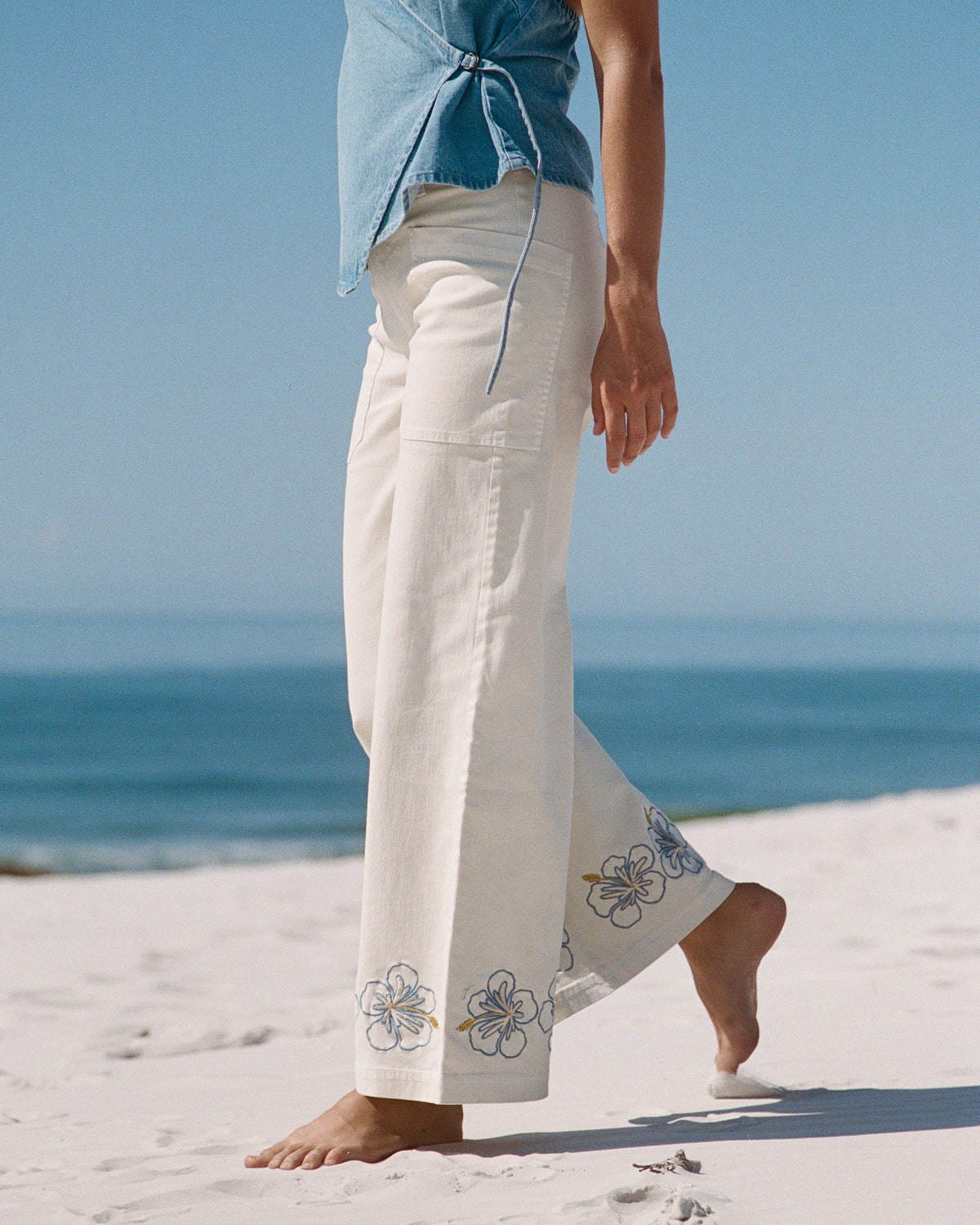 White cotton pants with embroidered hibiscus flowers at hem, worn on beach with blue tie-dye shirt.