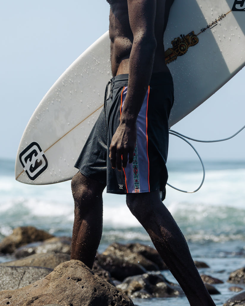 Black boardshorts with red and blue stripe detail, worn while holding a surfboard on a beach.