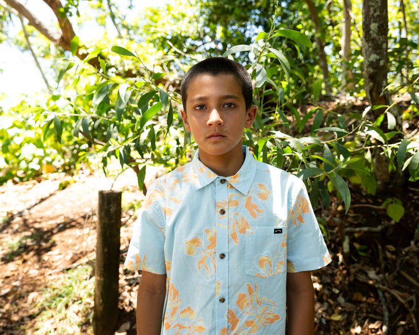 Young boy standing in a forested area wearing a light blue shirt with a floral pattern.