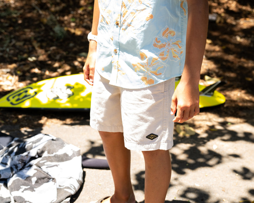 Person wearing a light blue shirt with gold designs and white shorts, standing outdoors with a yellow surfboard in the background.