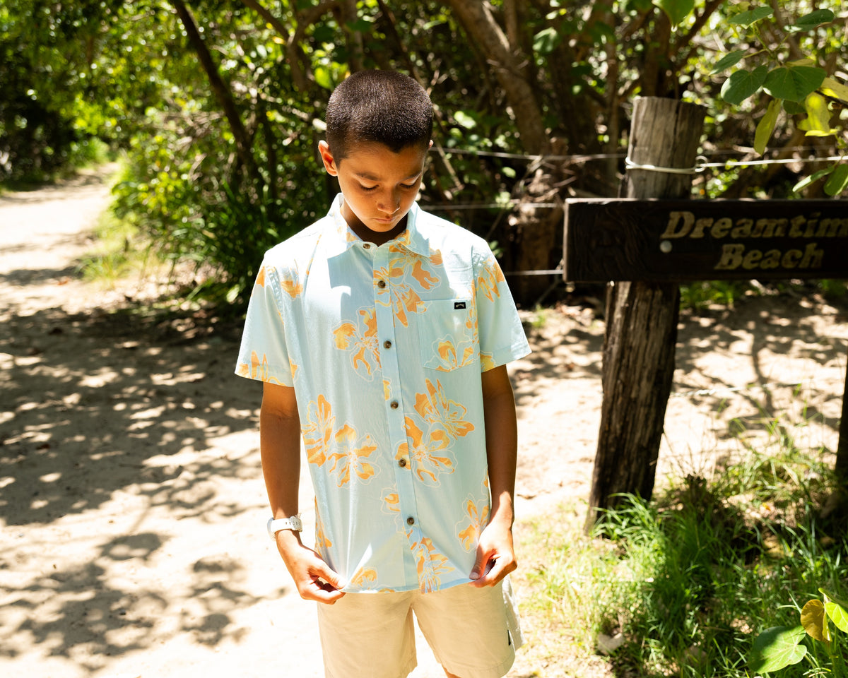 Person wearing a light blue floral shirt standing near a wooden signpost in a natural setting.