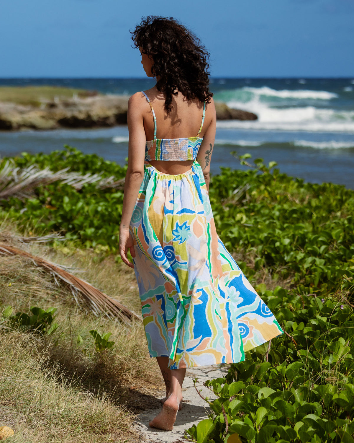 Sleeveless midi dress with blue, green, and yellow abstract print, worn on a beach with ocean background.