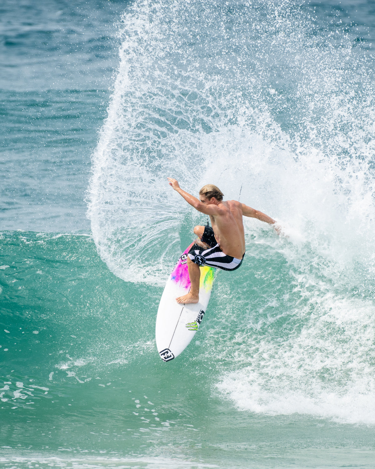 Shirtless surfer riding white and neon surfboard on large ocean wave.