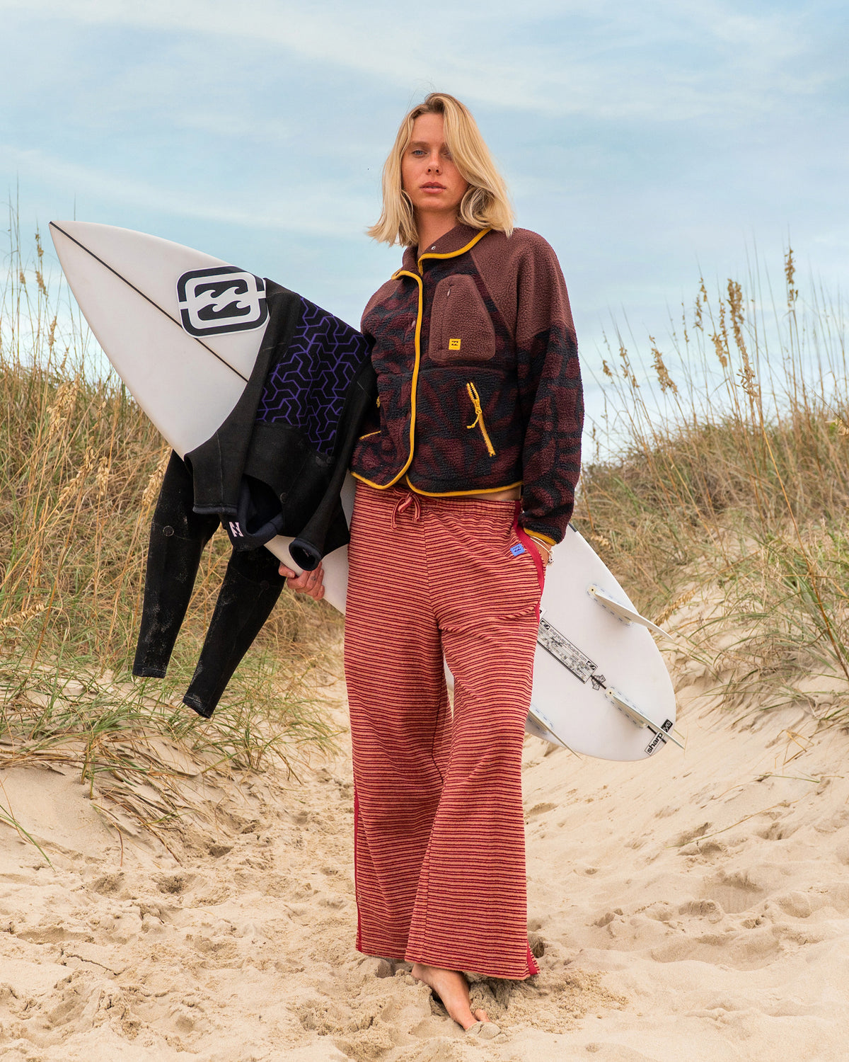Woman wearing rose brown horizontal striped terry pants with yellow trim at beach, holding surfboards.