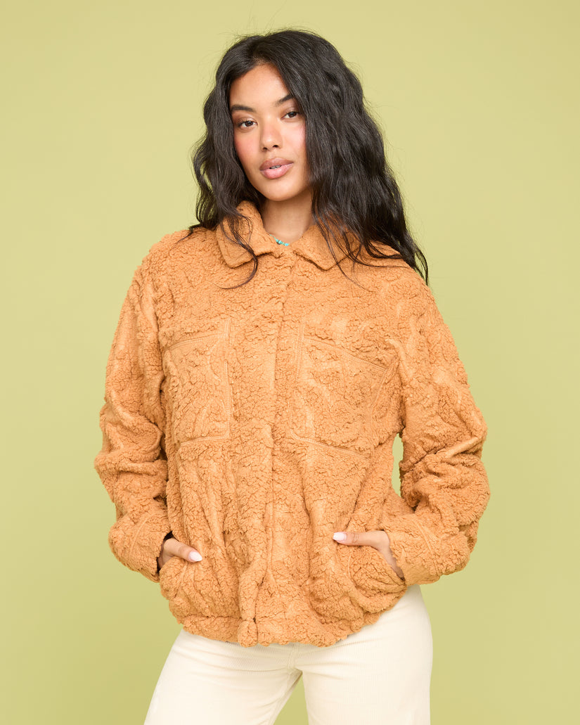 Camel-colored sherpa shacket with collar, button front, and patch pockets worn over cream pants.
