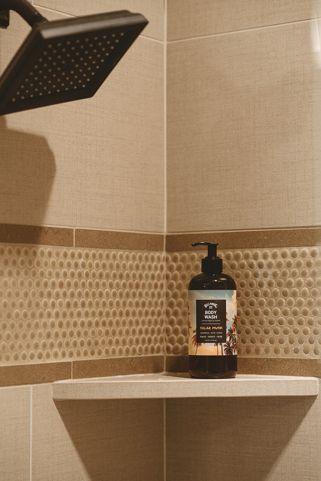 Black pump bottle of Solar Musk Body Wash displayed on a beige tiled bathroom shelf with showerhead visible above.