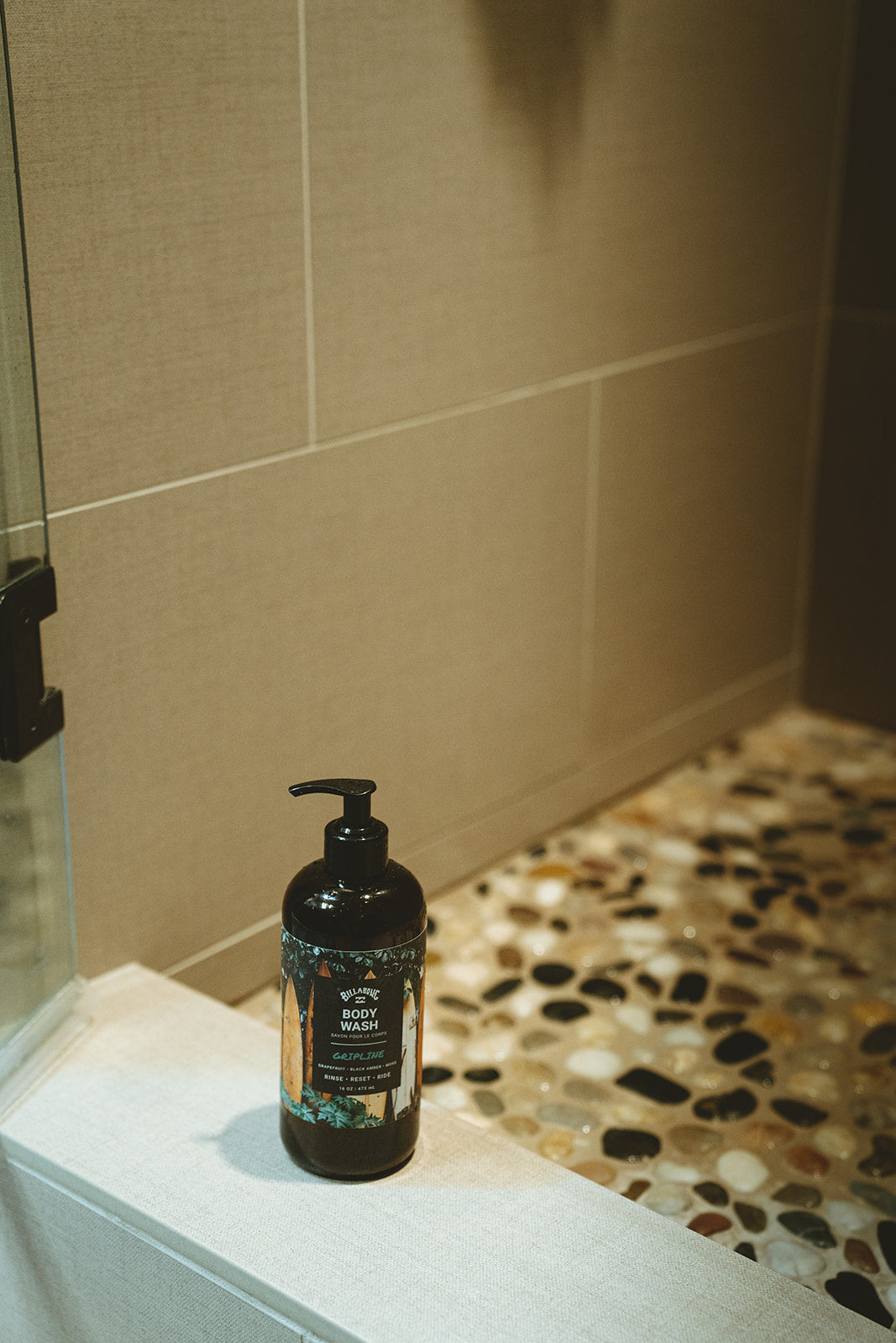 Black pump bottle of Gripline Body Wash on bathroom shelf with tiled walls and pebble floor visible.