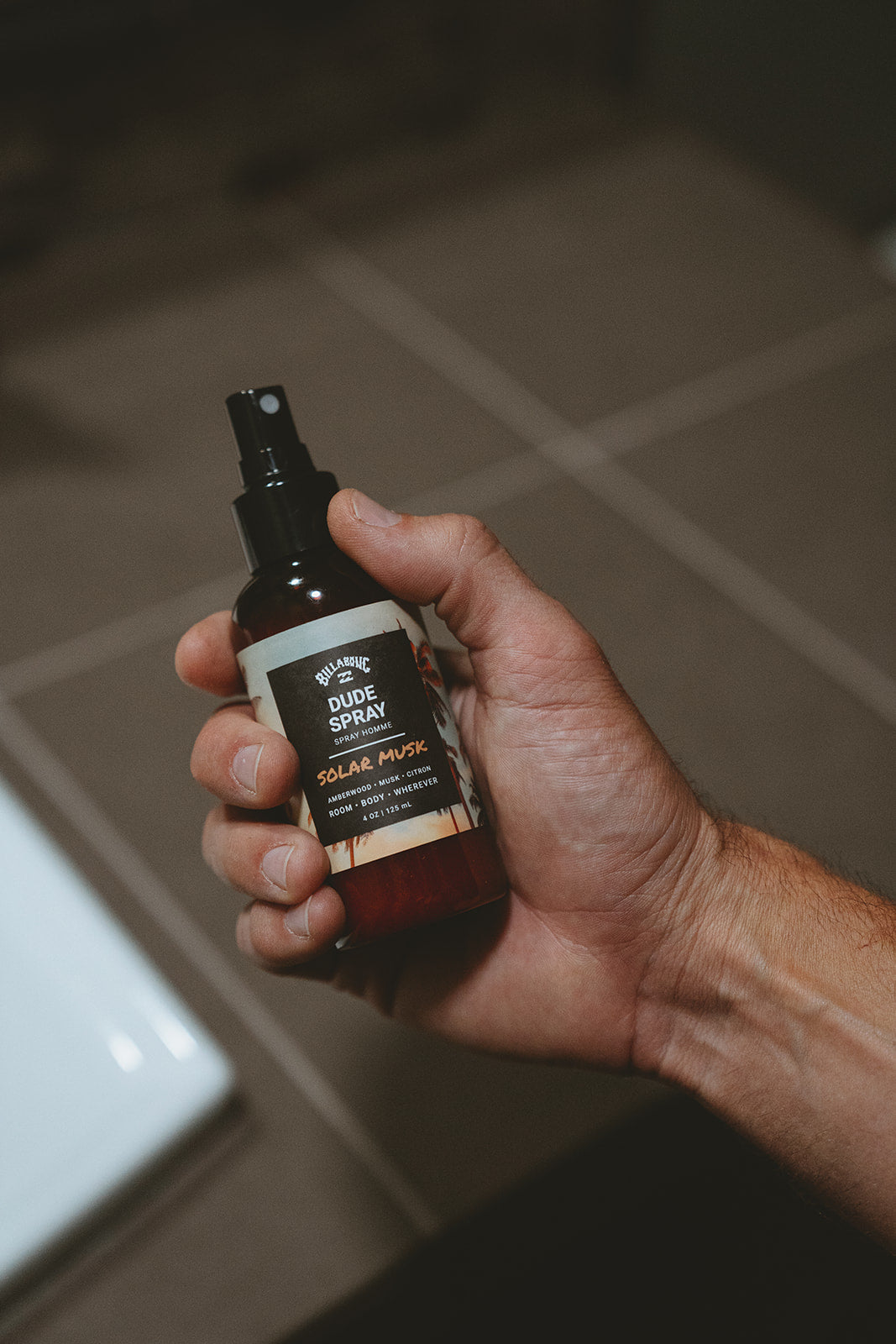 Dark glass spray bottle with black pump top and tan label reading "Dude Spray Solar Musk" held in hand.