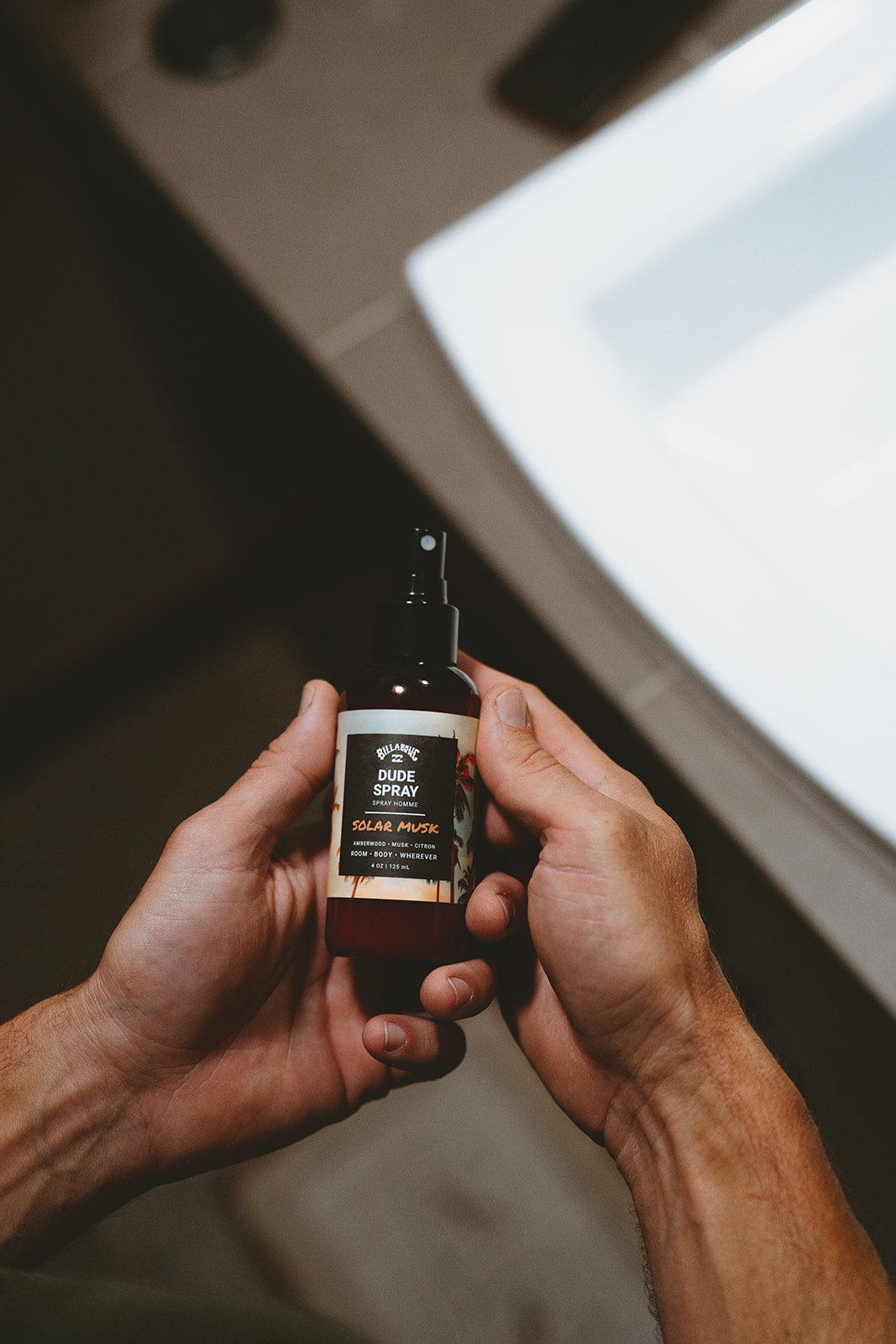 Two hands holding a dark spray bottle labeled "Solar Musk Dude Spray" with a black pump applicator.