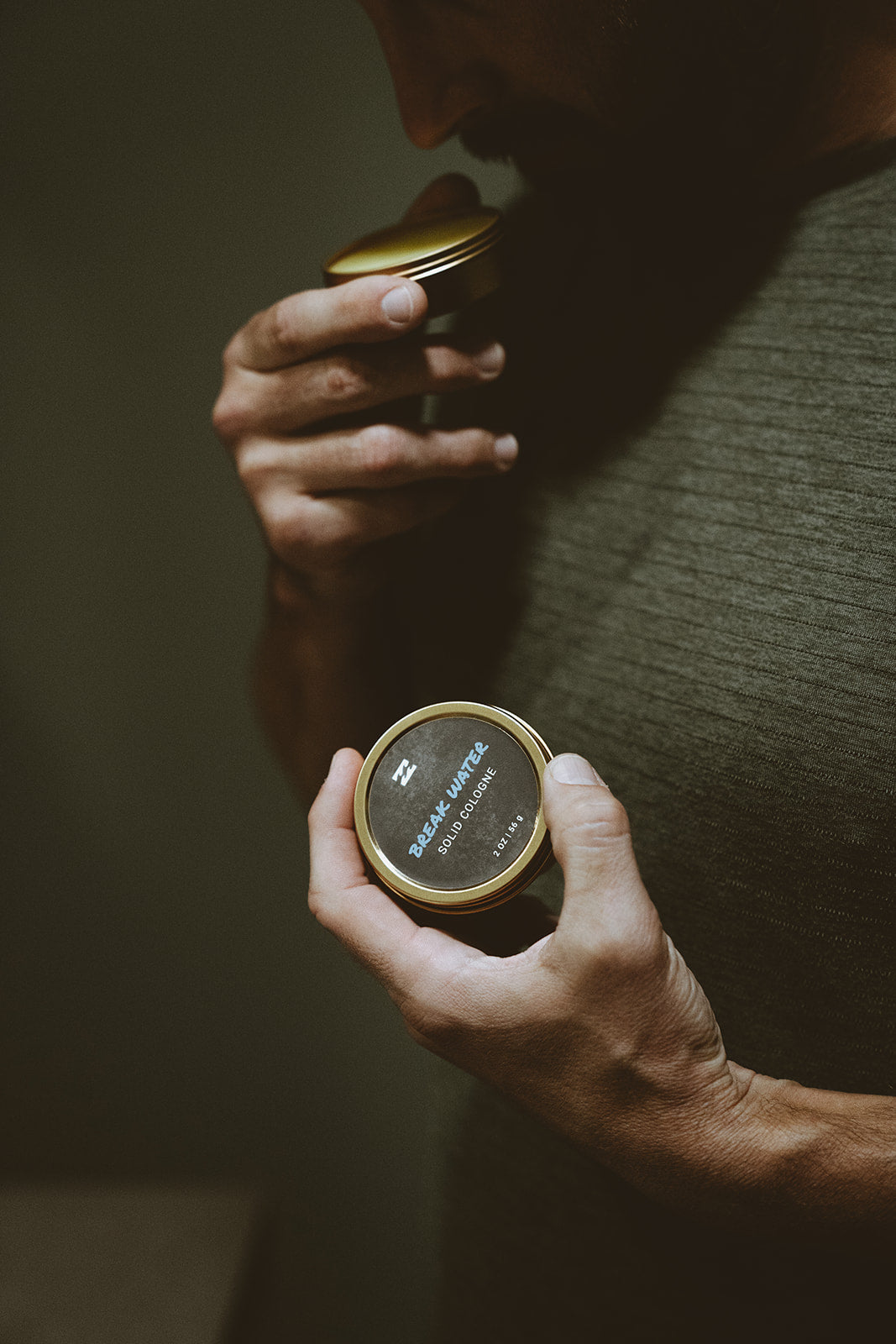 Hands holding a gold-rimmed tin container with dark label showing "Break Water Solid Cologne" text and product details.