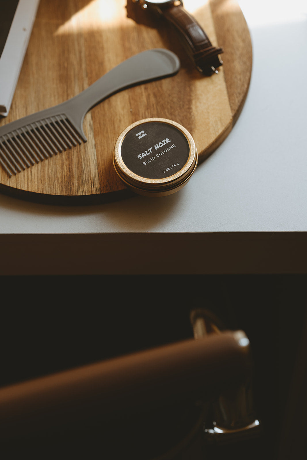 Round tin of Salt Noir solid cologne with gold rim on wooden surface next to gray comb and leather accessories.