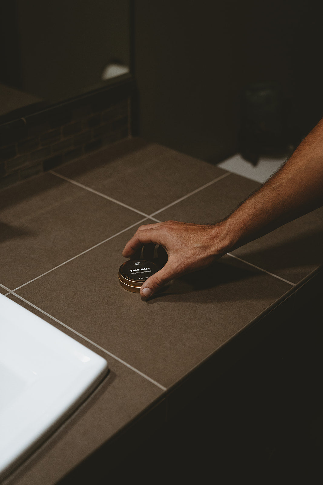 Hand holding a round black tin container with gold rim on a tiled bathroom countertop.