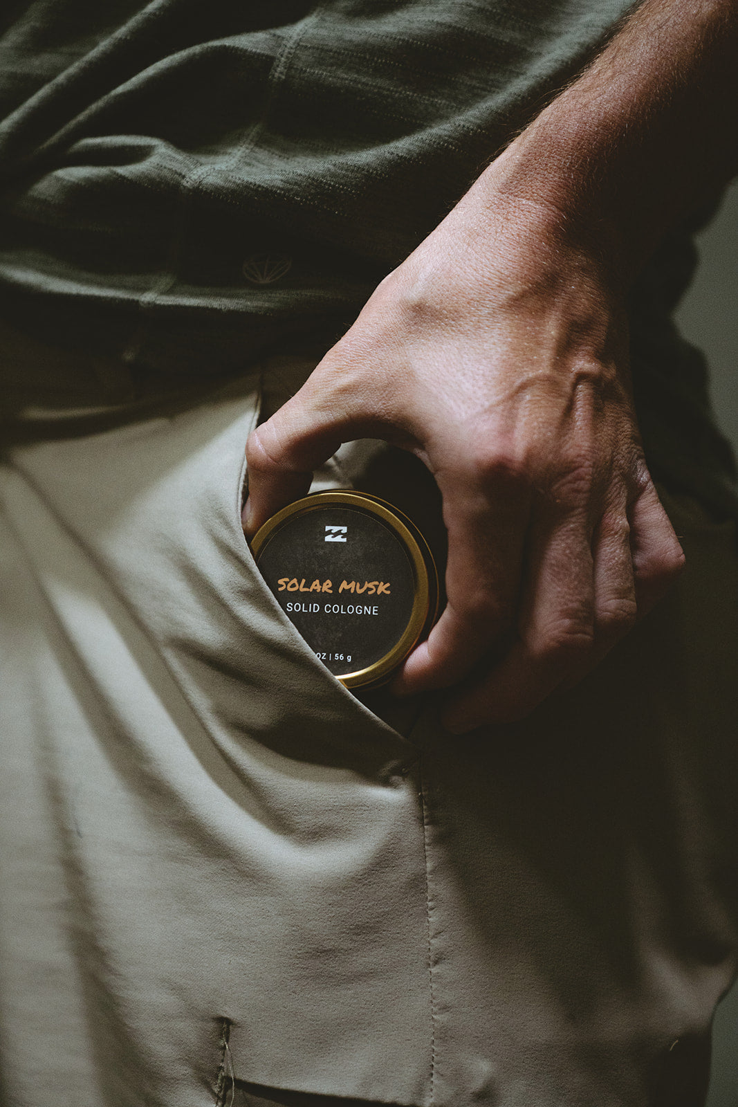 Hand holding round black tin with gold rim labeled "Solar Musk Solid Cologne" over khaki fabric.