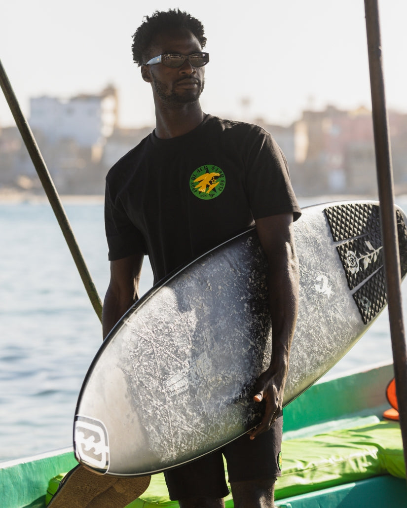 Black t-shirt with green and yellow circular logo on chest, worn with sunglasses while holding a surfboard.