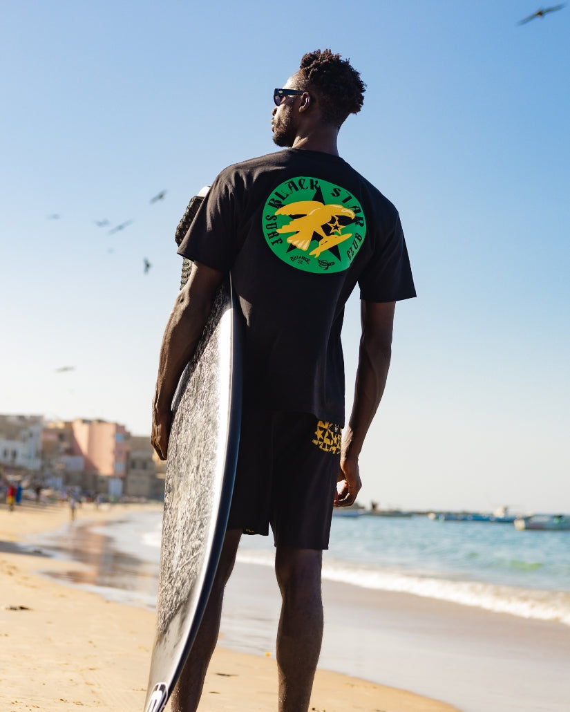 Black t-shirt with green and yellow star graphic on back, worn with surfboard at beach.