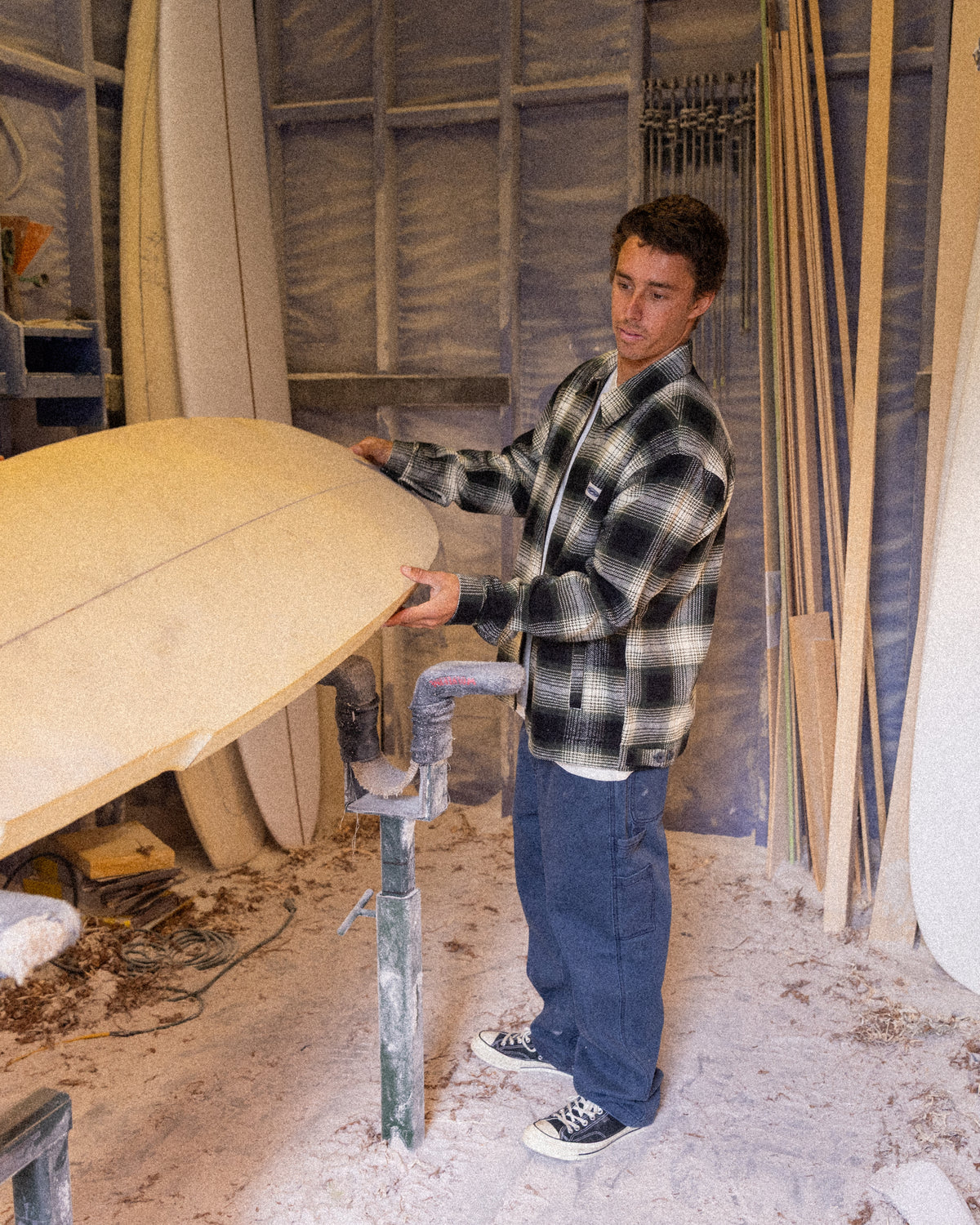 Man wearing black and white plaid jacket while working with surfboard in workshop.