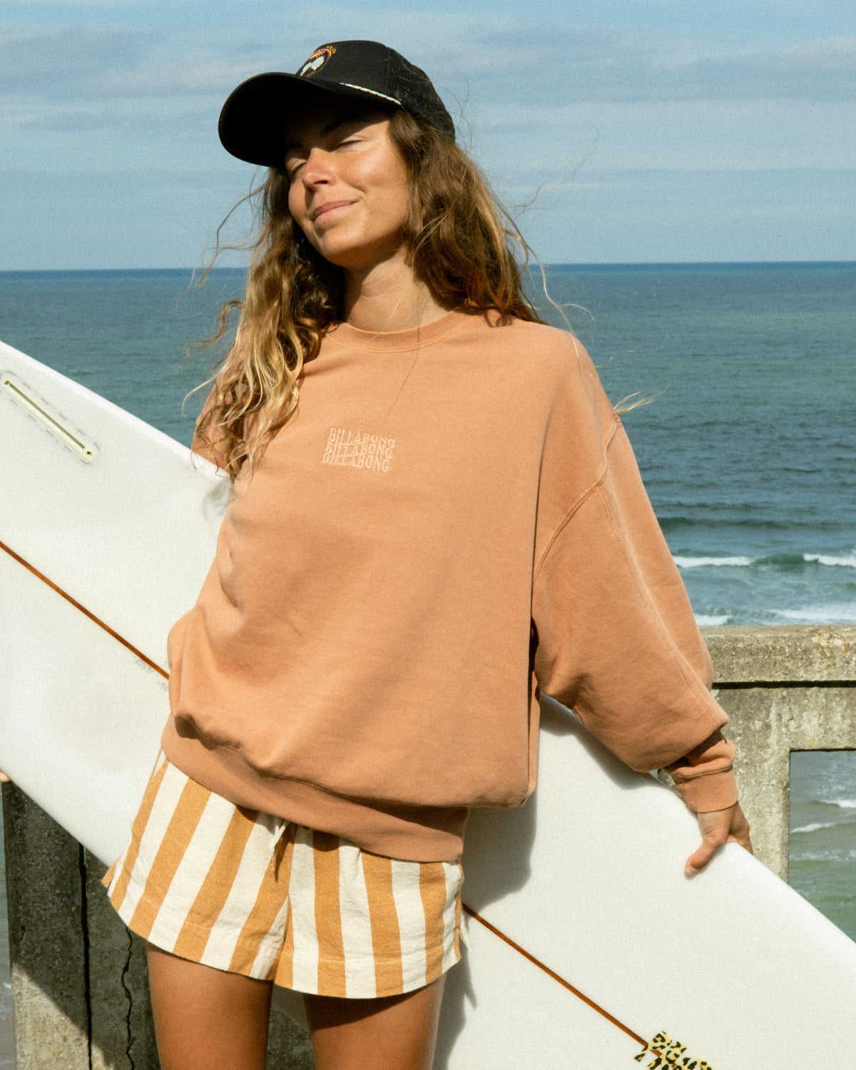 Caramel-colored crewneck sweatshirt with small chest graphic worn by person holding surfboard at beach.